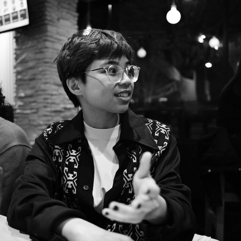 A black and white photo of Jasmine siting at a table in a restaurant, looking to their left and gesturing with their left hand. They appear to be in conversation with someone beside them. They are wearing glasses, a black buttoned shirt overtop of a white tee.