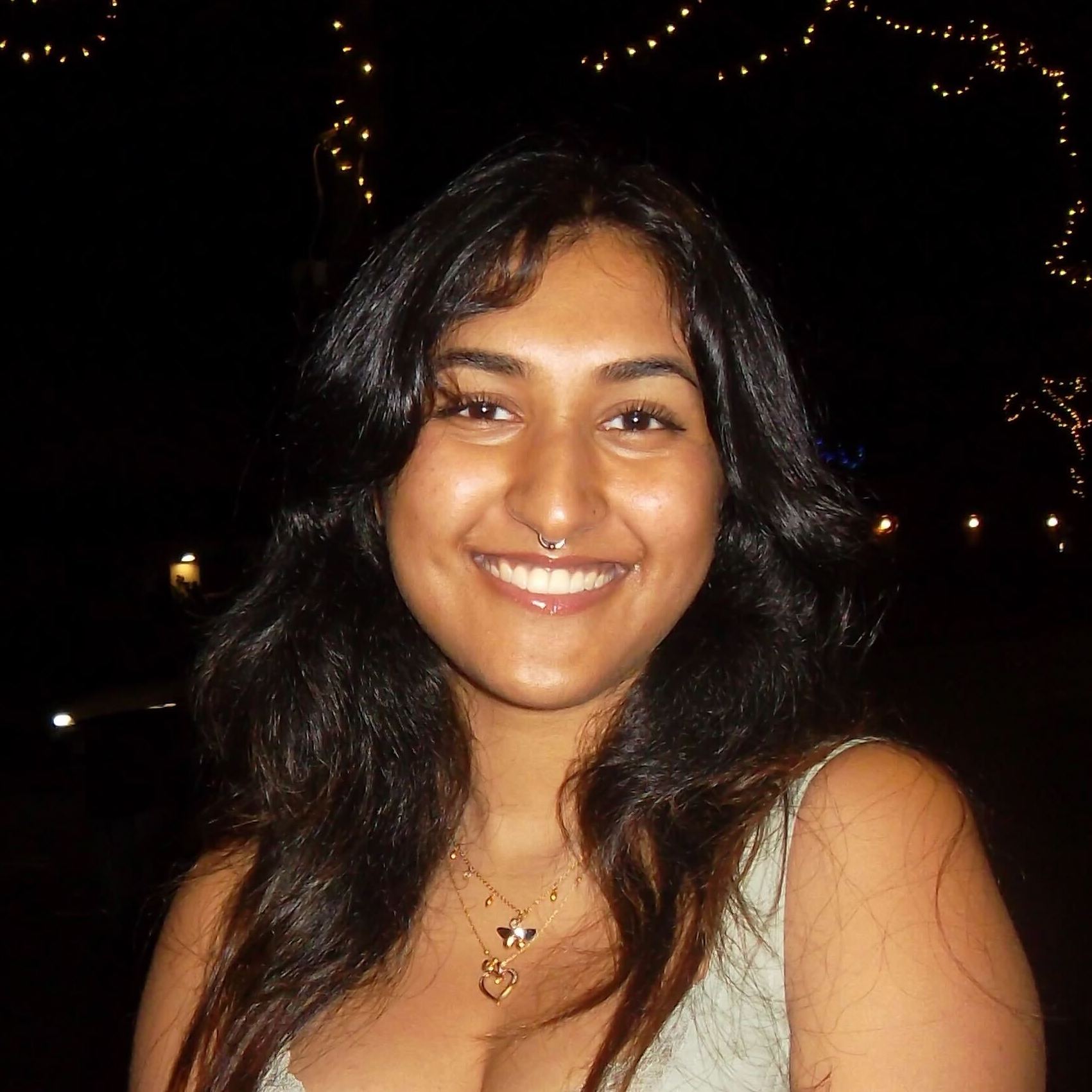Rea, a woman of colour with long dark-brown hair, smiles to the camera. The background is nearly black, but a few yellow spots of light from the trees can be seen.