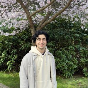 Darius, a man with dark hair wearing a cream hoodie and white jacket, stands smiling in front of a blossoming cherry tree.