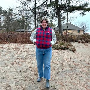Emily Ash Cutajar is photographed walking towards the camera atop sandy ground, with brush and sparse trees behind. She wears blue jeans and a grey sweater with a red and blue checkered vest overtop.