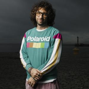 Gianluca Matarrese stands smiling at the camera, wearing glasses and a brightly coloured sweatshirt with the Polaroid logo on its front. Behind him is the darkened view of a harbour and sea vista.