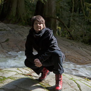 Liz Marshall, a person with short brown hair wearing an all-black outfit and red boots, is looking at the camera and crouching atop a large rock, with lush trees behind.