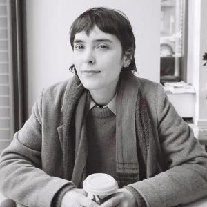 A black and white photograph of Sara Wylie, a white woman with short brown hair wearing a scarf, knit sweater and coat and holding a to-go coffee cup. Sarah looks at the camera with a small smile, and appears to be sitting in a cafe.