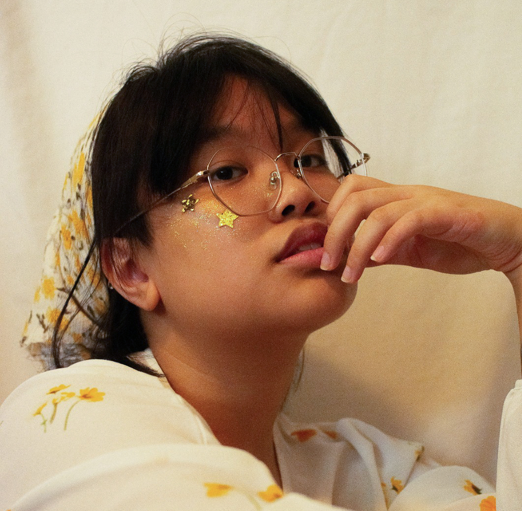 jas calcitas is pictured in front of a cream-coloured backdrop, their black hair tied back in a white and yellow scarf that matches their shirt, which has yellow flowers printed on it. They have sparkly stars on their cheeks, and are wearing wire-framed glasses.