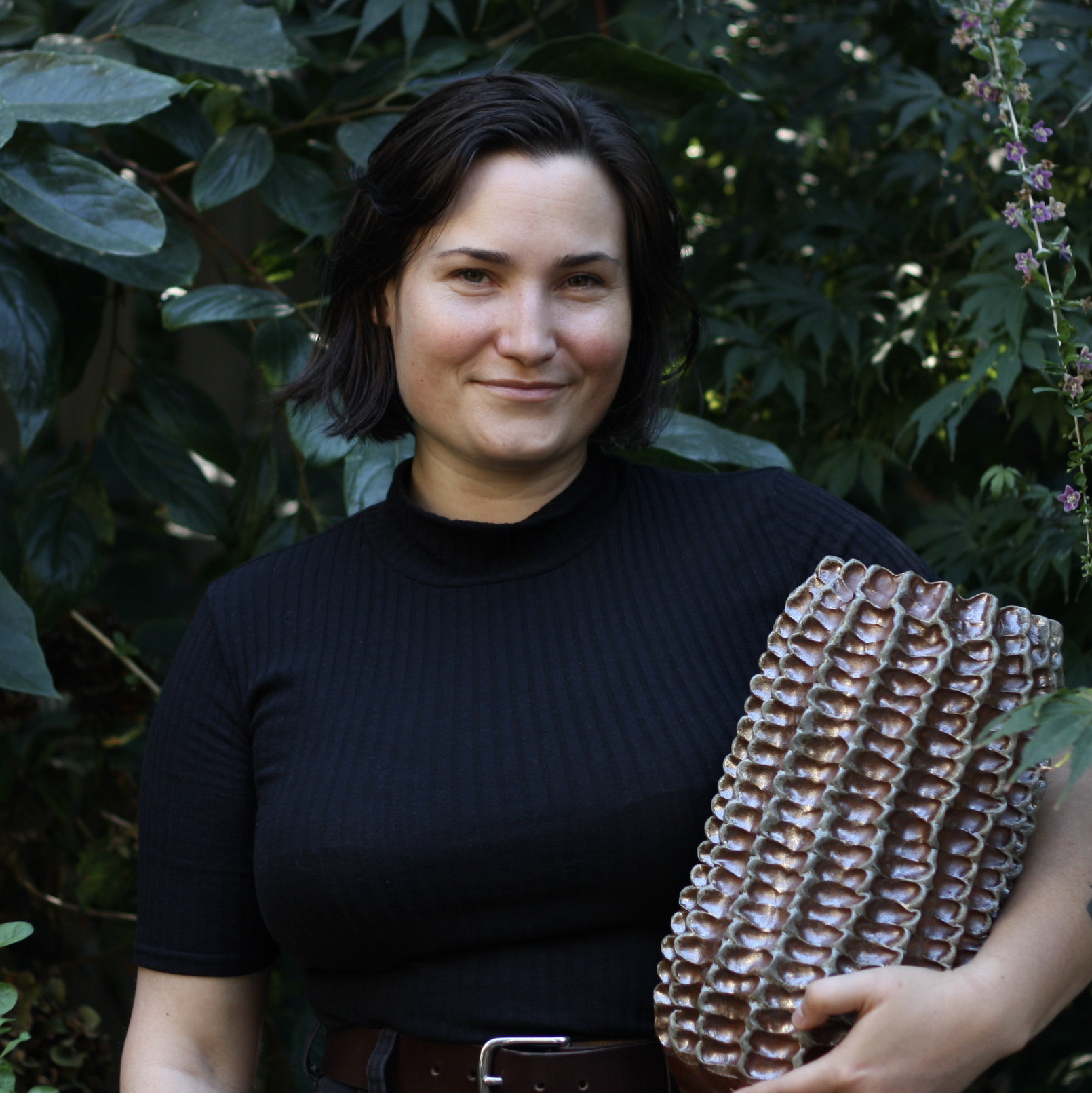 Brit Bachmann stands, smiling warmly with her mouth closed, in front of a deep and lush collection of greenery. She is wearing a black mock beck top with sleeves ending at the elbow, and a dark brown leather belt. In her left arm she is holding a large ceramic vessel, which is covered in rows of pinched textures and has a bronzy hue.