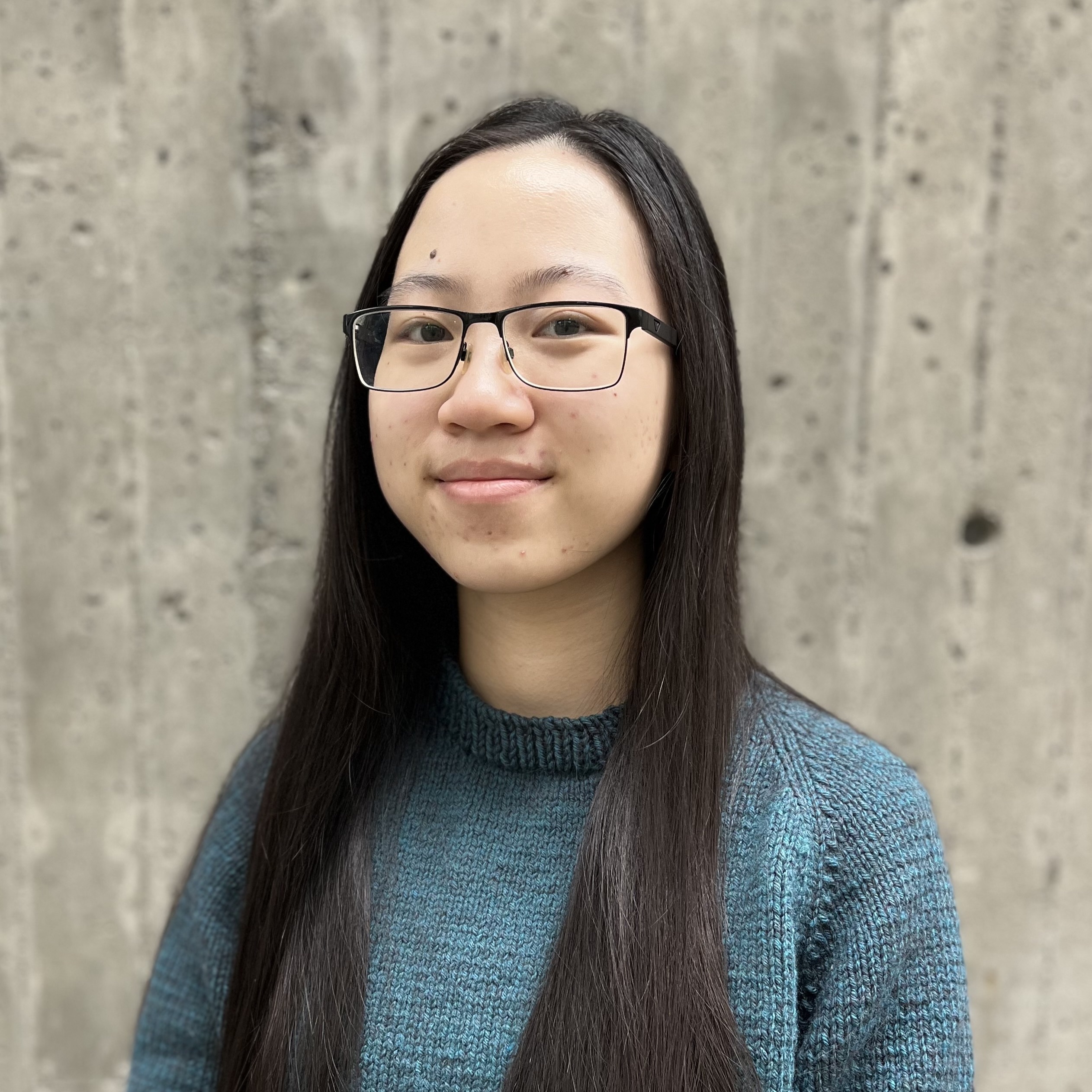 Erica stands in front of a grey cement backdrop. She wears a blue knit sweater, visible from the shoulders up, and her long, straight black hair falls down past the photo frame. Erica is wearing a pair of black glasses with rectangular frames, and has a slight smile on her face.