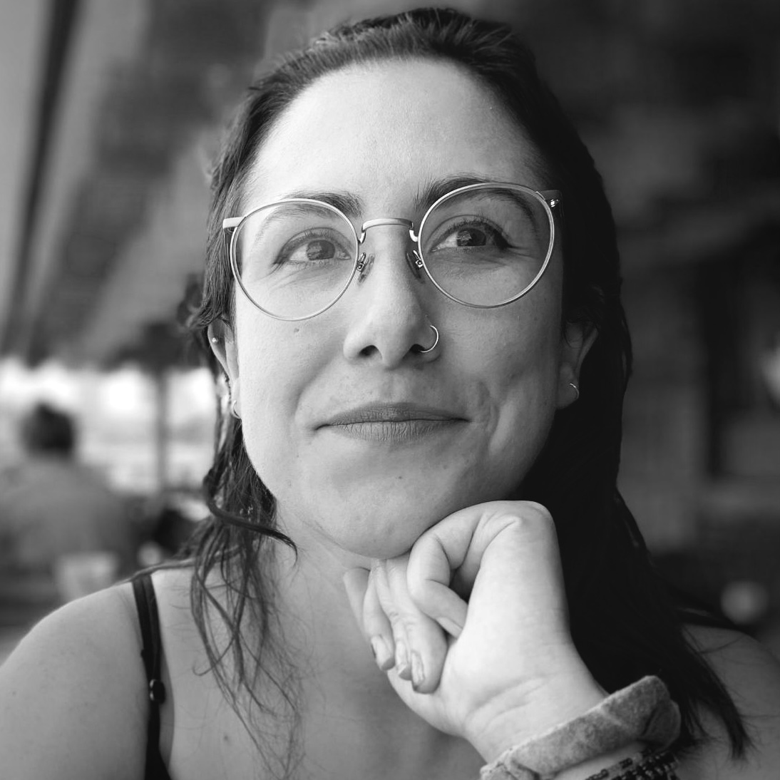 Black and white photograph of Laura Arboleda, who is smiling with her mouth closed and her hand comfortably resting underneath her chin. She wears thin-framed, circular classes and her dark hair is pushed away from her face, with a few strands hanging down by her shoulder. The background is slightly blurry, but she appears to be on an outdoor covered patio of some kind.