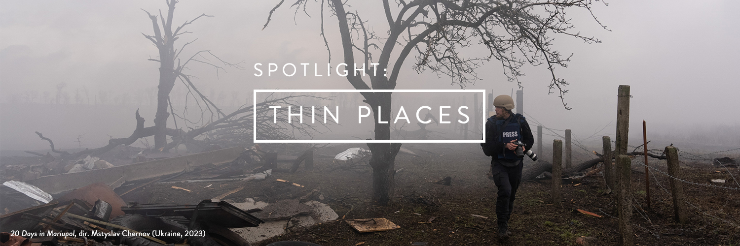 Film still showing a labelled journalist walking through a misty or smoky path of destruction, burned trees, holding a camera. White text and a white box that reads Thin Places. Film still from 20 Days in Mariupol, 2023