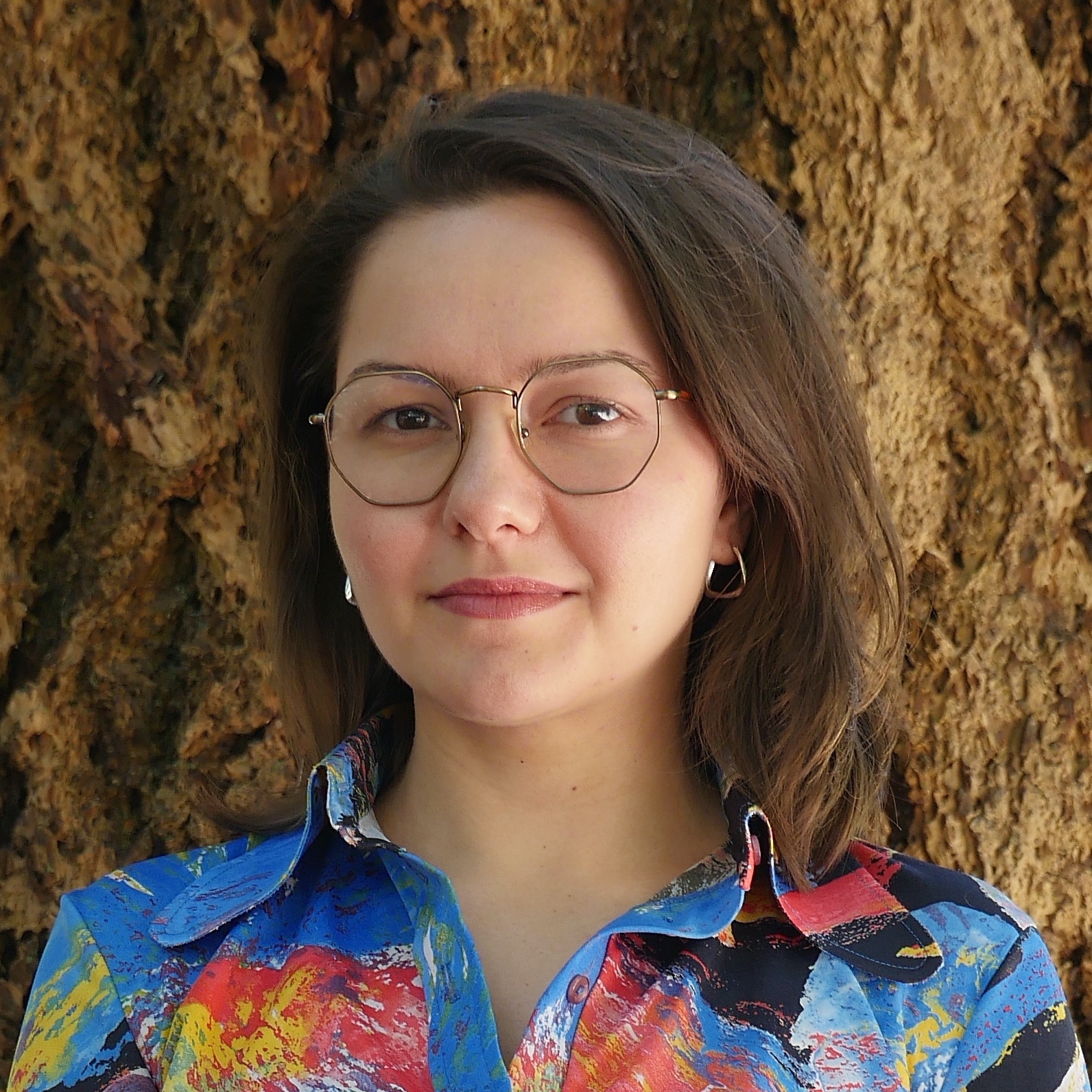 Özgün is pictured from the shoulders up in front of the trunk of a tree. She is looking at the camera and smiling with her mouth closed. She wears glasses and a brightly coloured shirt.