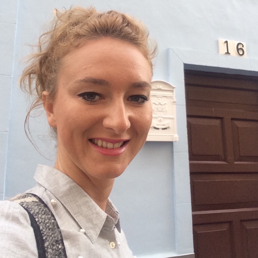 Jelena, a white woman with blonde hair pulled up in a messy bun, smiles at the camera while standing in front of a pale blue wall and brown garage door.