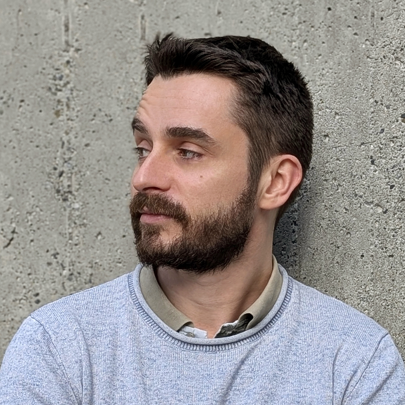 Photo of a man with brown hair and a brown beard, wearing an icy blue sweater and leaning against a concrete wall.