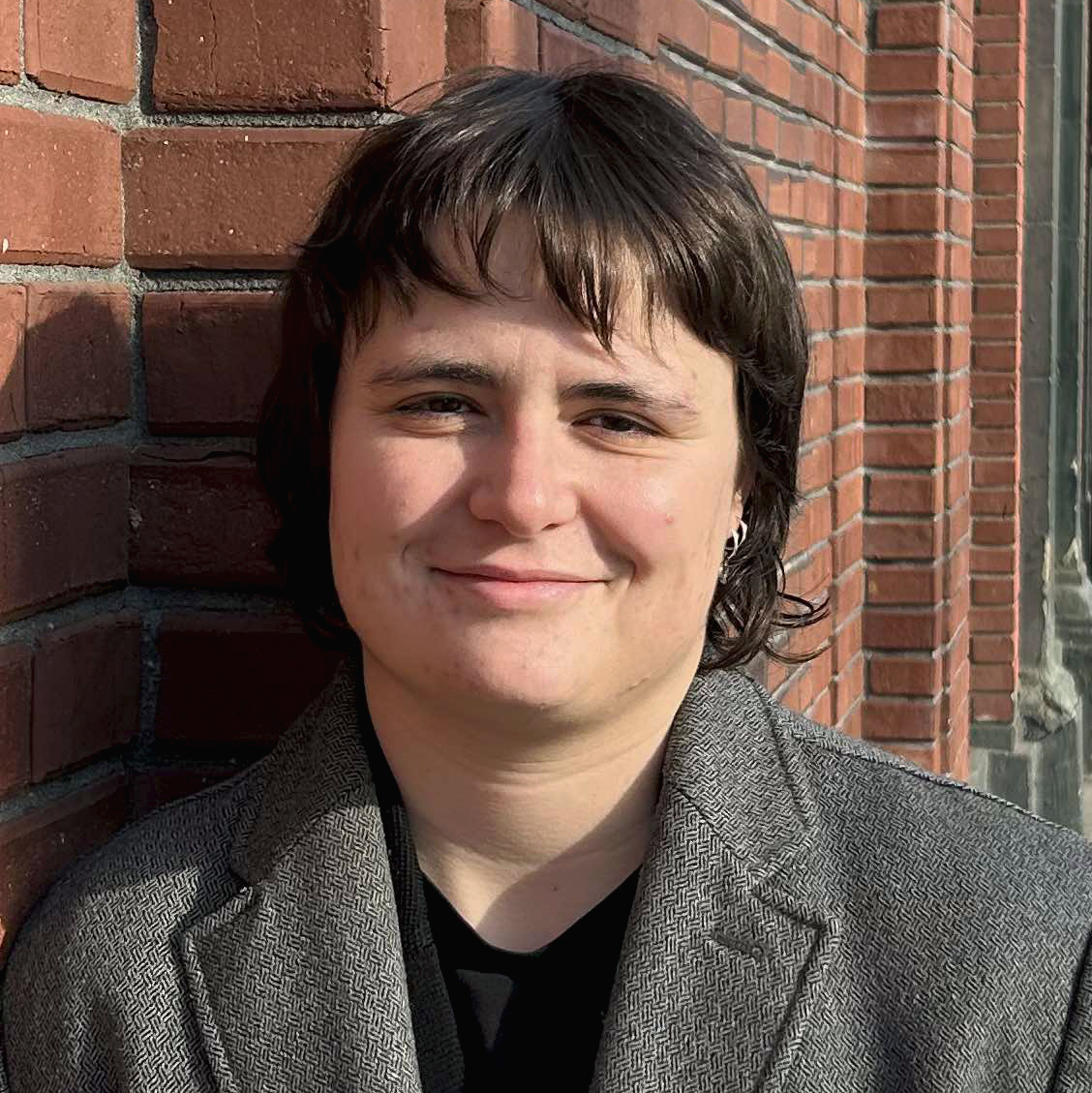 A person with short brown hair and a grey blazer leans against a brick wall, with sun shining on their face.