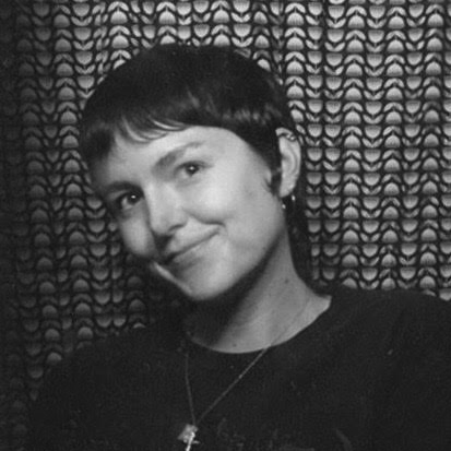 Black and white photo of a person with a pixie haircut, tilted head and mischievous smile.