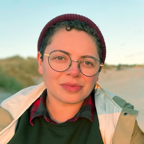 Headshot of a person on the beach, waring a cranberry toque and a pistachio green jacket.