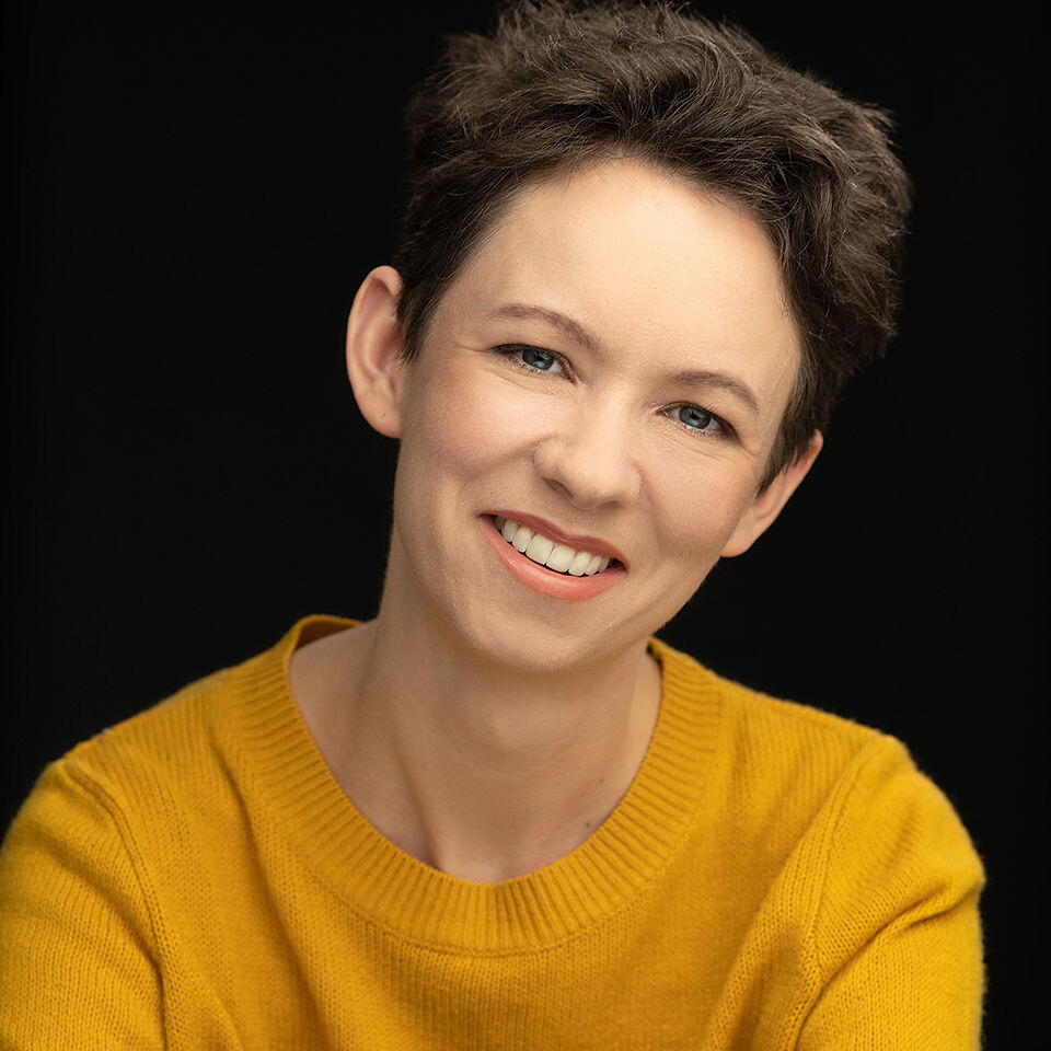 A person with short brown hair smiling warmly at the camera, wearing a mustard yellow sweater against a black background.