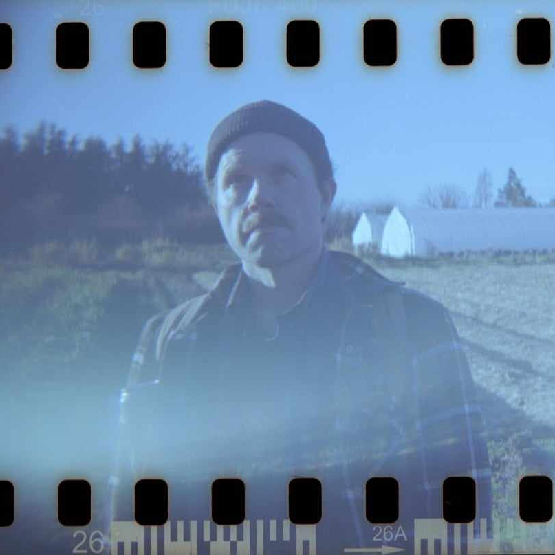 A slightly faded, filmstrip-style photo of a man wearing a dark beanie and jacket standing outdoors near a field and a long white greenhouse or barn, with trees in the background under a clear sky.