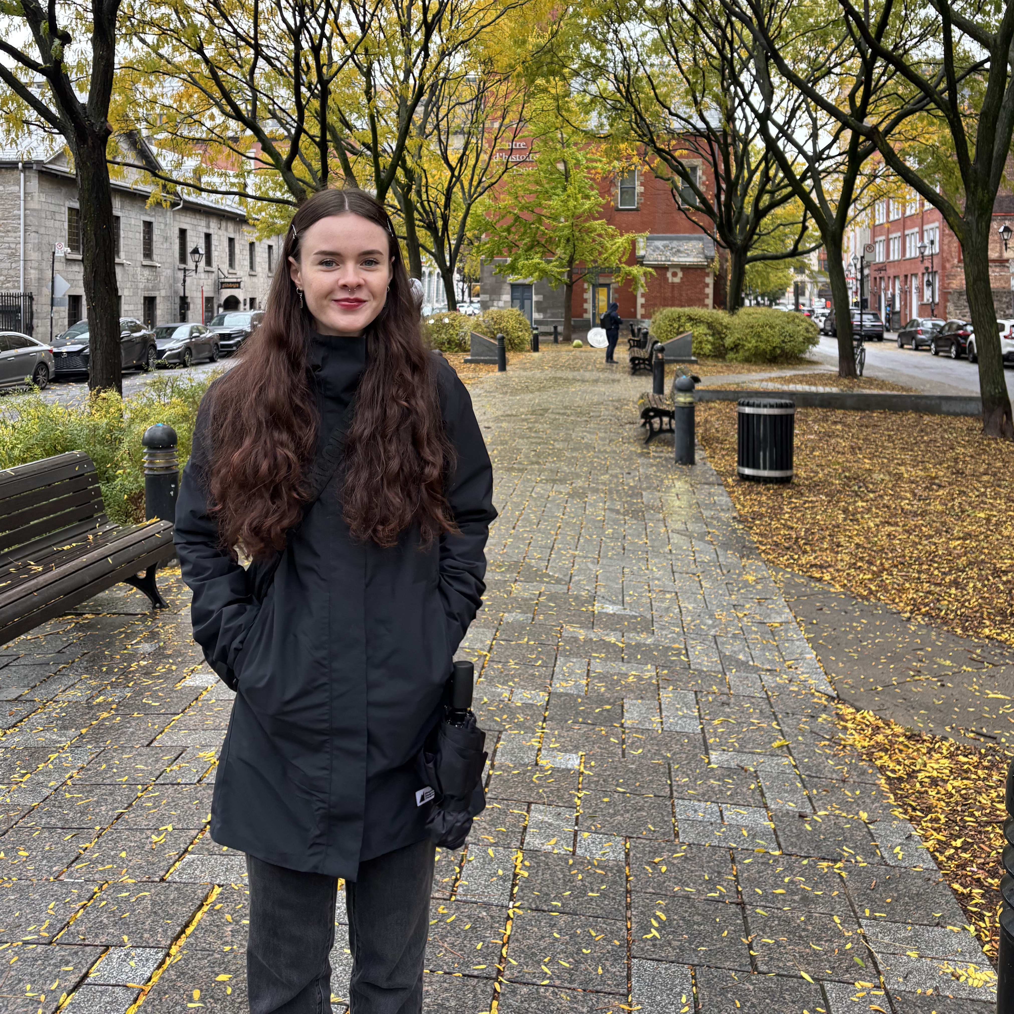A woman with long wavy brown hair stands on a tree-lined city sidewalk in autumn, wearing a black coat with her hands in the pockets. Fallen yellow leaves scatter across the pavement, with benches, parked cars, and stone and brick buildings in the background.