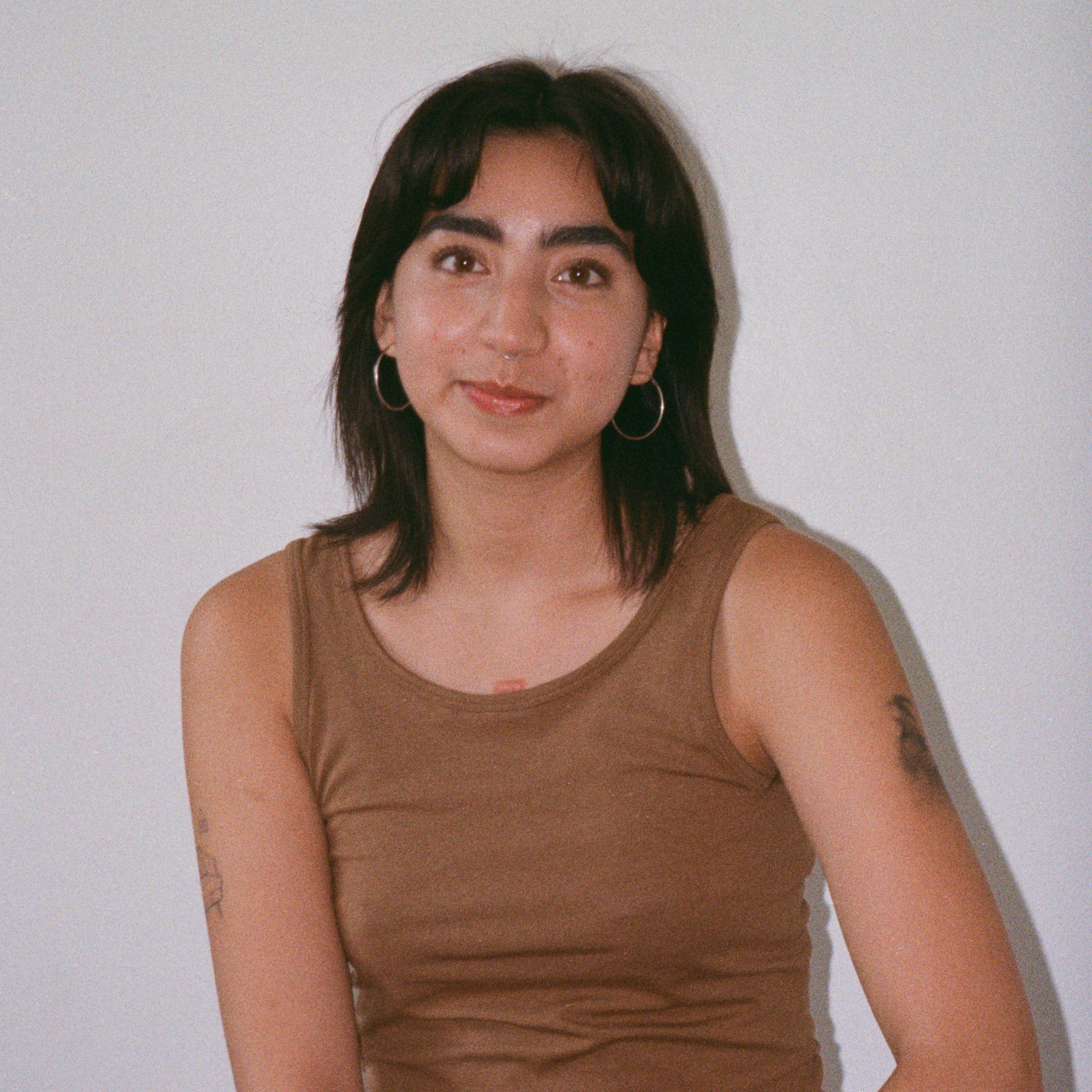 A person with short dark hair and bangs sits against a plain light-colored wall, wearing a brown sleeveless top and hoop earrings. They have a subtle smile and visible arm tattoos, and the photo has a soft, slightly grainy look.