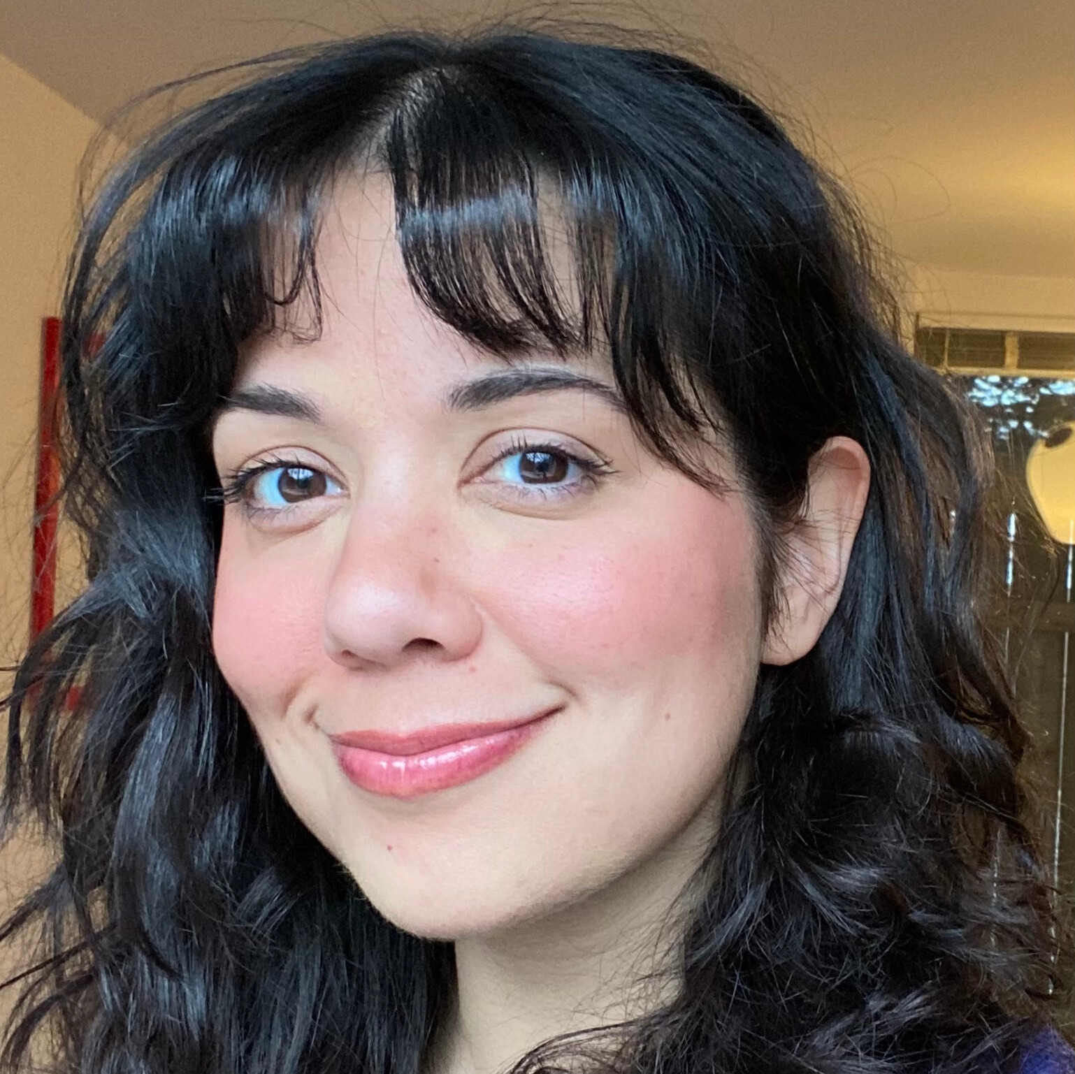 A person with wavy dark hair and short bangs smiles softly at the camera indoors. They have rosy cheeks and natural makeup, and are wearing a purple sweater over a red shirt. Warm indoor lighting and a window are visible in the background.
