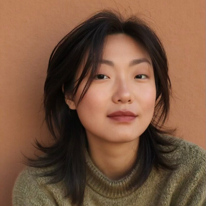A young Asian woman with shoulder-length dark hair and natural makeup, wearing a cozy olive-green knit sweater, looks confidently into the camera with a neutral expression. She is posed against a warm, tan-colored background.