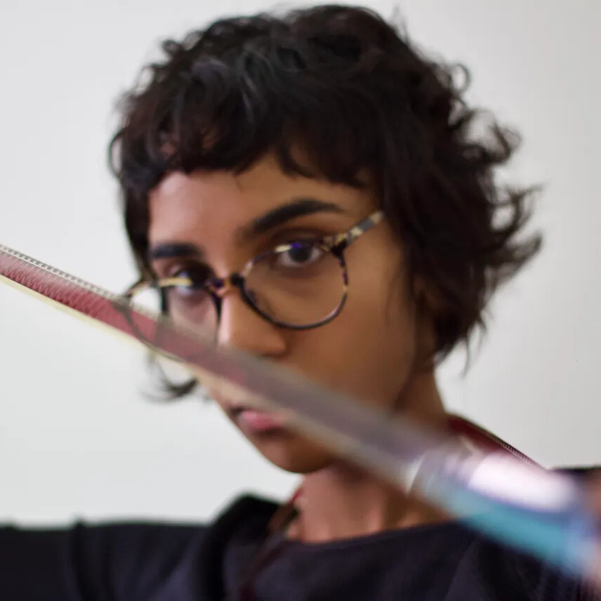 A person with short, curly dark hair and round glasses stretches a piece of 16mm film toward the camera. The 16mm runs diagonally across the frame and is slightly blurred in the foreground, while the person looks intently past it against a plain background.