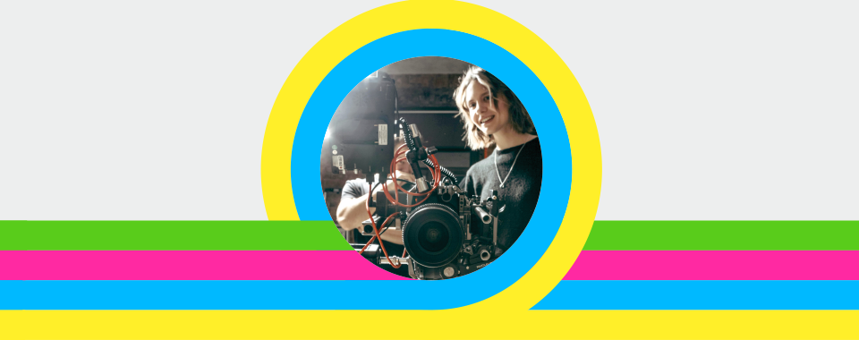 A woman smiles behind a large camera, encircled by vibrant rings of yellow and blue. Background features horizontal green, pink, and blue stripes.