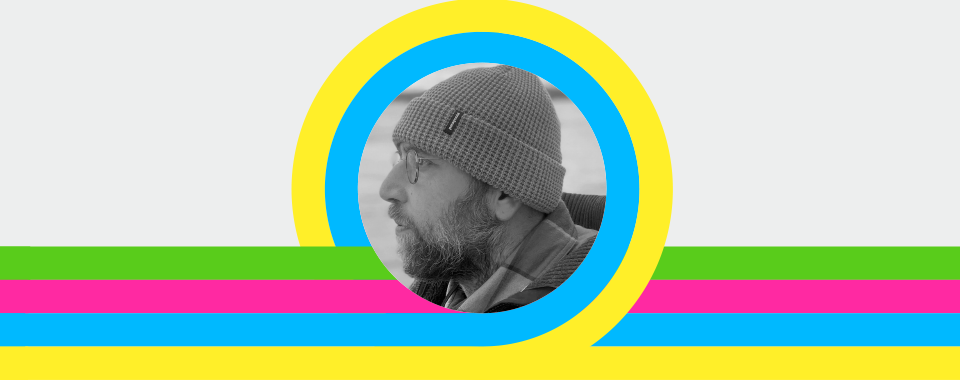 Black and white side profile portrait of a bearded man wearing glasses and a knit beanie, framed in a circular crop with bold yellow and blue rings, over a gray background with horizontal green, pink, and blue stripes; modern graphic design banner style.