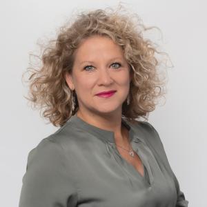 Julie Roy, a woman with curly shoulder-length blonde hair and wearing pink lipstick, smiles and looks with one shoulder angled closer to the camera against a white background. She is wearing a shiny grey blouse and silver jewelry.