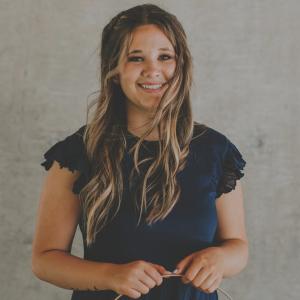 Anna Hetherington, a woman with long brownish-blonde hair wearing a navy blue dress, stands against a grey stone backdrop and smiles at the camera.