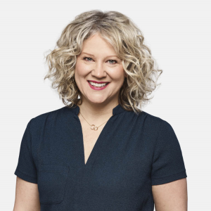 Julie Roy, a white woman with short curly blonde hair wearing a black shirt and gold necklace, smiles and stands against a white backdrop.