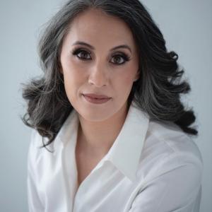 Woman with shoulder-length wavy gray hair and light makeup wearing a white collared shirt, looking at the camera against a plain light background.