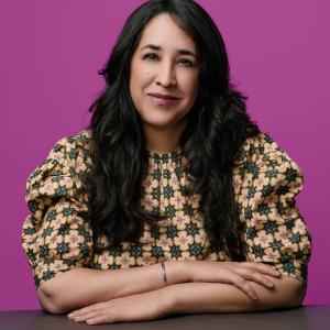 Woman with long dark hair sitting at a table, wearing a patterned blouse, against a bright pink background.
