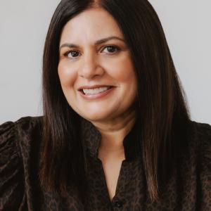Smiling woman with straight dark hair wearing a dark patterned blouse against a light background.
