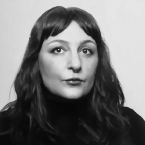Black and white portrait of a woman with long, wavy hair wearing a dark turtleneck, looking slightly off-camera with a neutral expression.