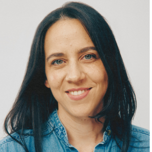 Woman with long dark hair and blue eyes smiles at the camera, wearing a blue denim shirt against a plain light background.