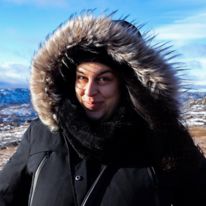 A person in a black coat with a fur-trimmed hood smiles warmly in a snowy landscape. The bright blue sky adds contrast and a cheerful tone.