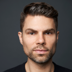 Portrait of a person with short, dark brown hair and a trimmed beard, looking directly at the camera against a dark gray background. The expression is neutral.
