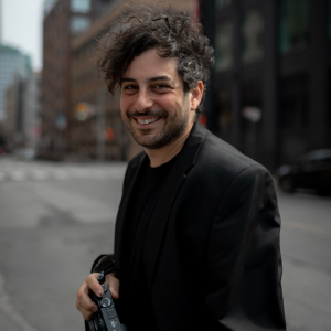 Man with curly hair and stubble smiling, holding a camera on a city street. He wears a black blazer, conveying a relaxed, cheerful vibe.