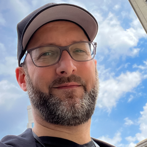 A person with a beard and glasses wearing a cap smiles slightly at the camera. The background features a blue sky with fluffy clouds.