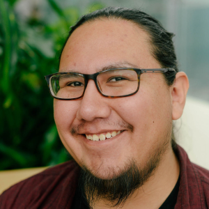 A person with glasses and a beard smiles warmly, set against a backdrop of green plants. The atmosphere is friendly and inviting.