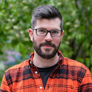 A man with short dark hair, a beard, and glasses, wearing an orange plaid shirt, smiles gently with lush green foliage in the background.