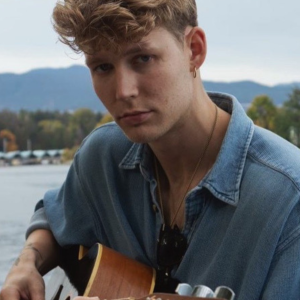 A person with wavy hair in a denim shirt plays guitar by a lake, with mountains and trees in the background. The mood is calm and introspective.