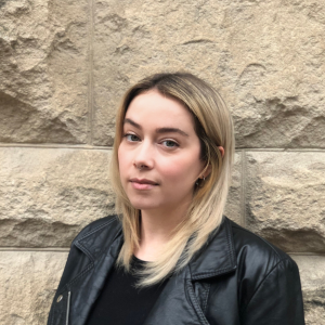 Woman with blonde hair in a black leather jacket stands against a textured stone wall, gazing confidently at the camera.
