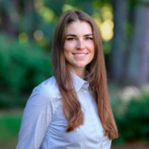 Smiling woman with long brown hair in a striped shirt stands outdoors. Background is blurred green foliage, creating a calm, natural setting.